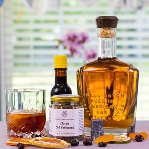 May include: A glass bottle of amber liquid with a wooden stopper, a small jar labeled Kristin's Koncoctions Classic Old Fashioned, a small bottle with a yellow cap, a glass of dark liquid with ice, and dried fruit slices and dark berries.