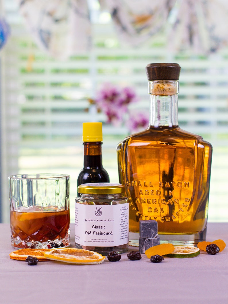 May include: A glass bottle of amber liquid with a wooden stopper, a small jar labeled Kristin's Koncoctions Classic Old Fashioned, a small bottle with a yellow cap, a glass of dark liquid with ice, and dried fruit slices and dark berries.