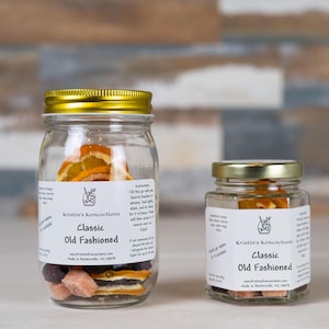 May include: Two clear glass jars with gold lids containing dehydrated fruit slices and sugar cubes for a Classic Old Fashioned cocktail. Each jar features a white label with the text Kristin's Koncoctions, Classic Old Fashioned, and instructions.