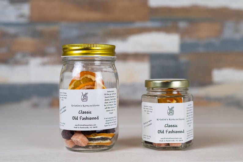 May include: Two clear glass jars with gold lids containing dehydrated fruit slices and sugar cubes for a Classic Old Fashioned cocktail. Each jar features a white label with the text Kristin's Koncoctions, Classic Old Fashioned, and instructions.