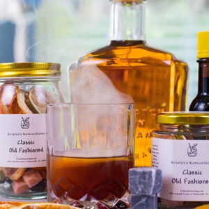 May include: A glass of amber liquid with rising smoke, flanked by two glass jars labeled Kristin's Koncoctions Classic Old Fashioned containing dried fruit and sugar cubes, a large amber glass bottle, a small yellow-capped bottle, and dark cherries.