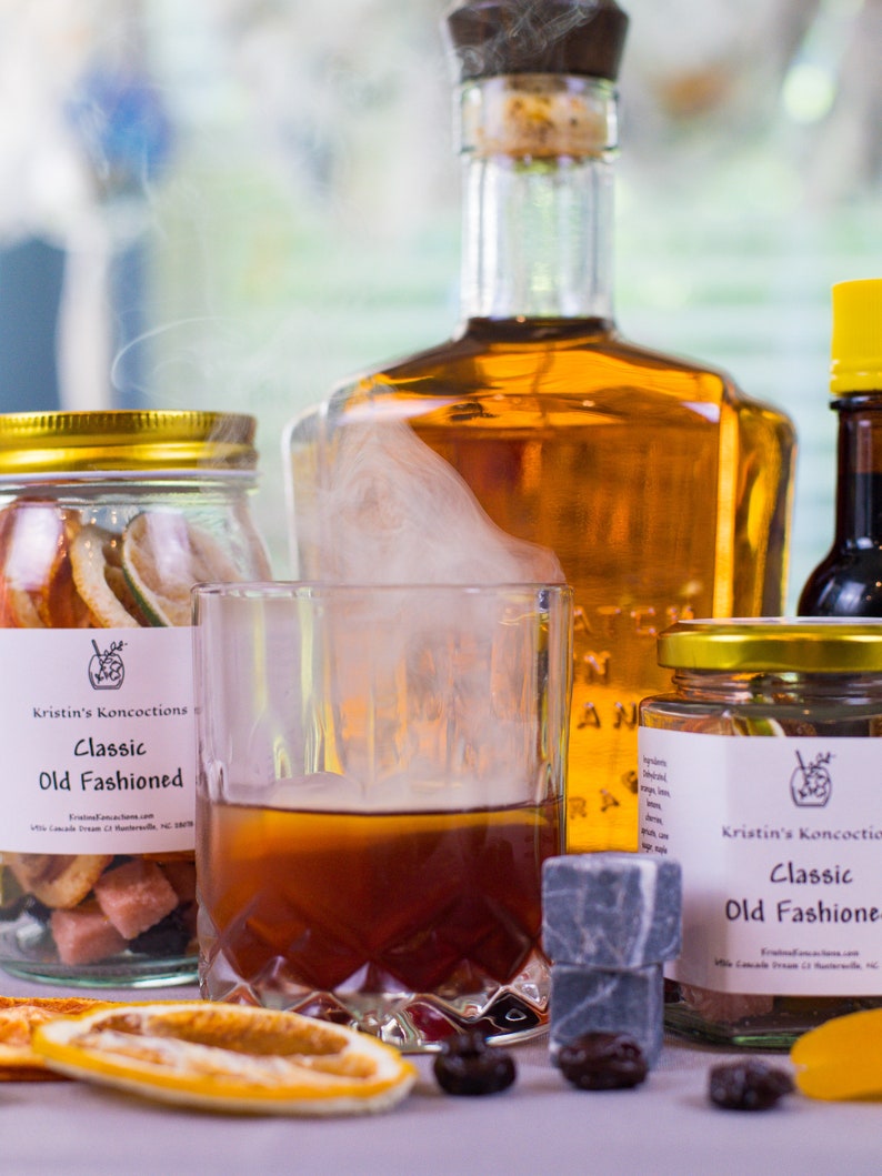 May include: A glass of amber liquid with rising smoke, flanked by two glass jars labeled Kristin's Koncoctions Classic Old Fashioned containing dried fruit and sugar cubes, a large amber glass bottle, a small yellow-capped bottle, and dark cherries.