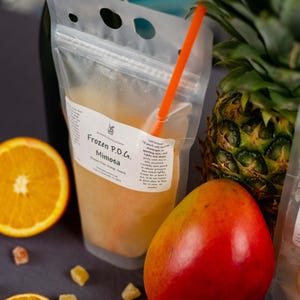 May include: A clear plastic pouch labeled "Frozen P.O.G. Mimosa" with an orange straw, next to a sliced orange, a mango, and a pineapple. The pouch contains a yellow-orange liquid. Small pieces of candied fruit are scattered around the pouch.