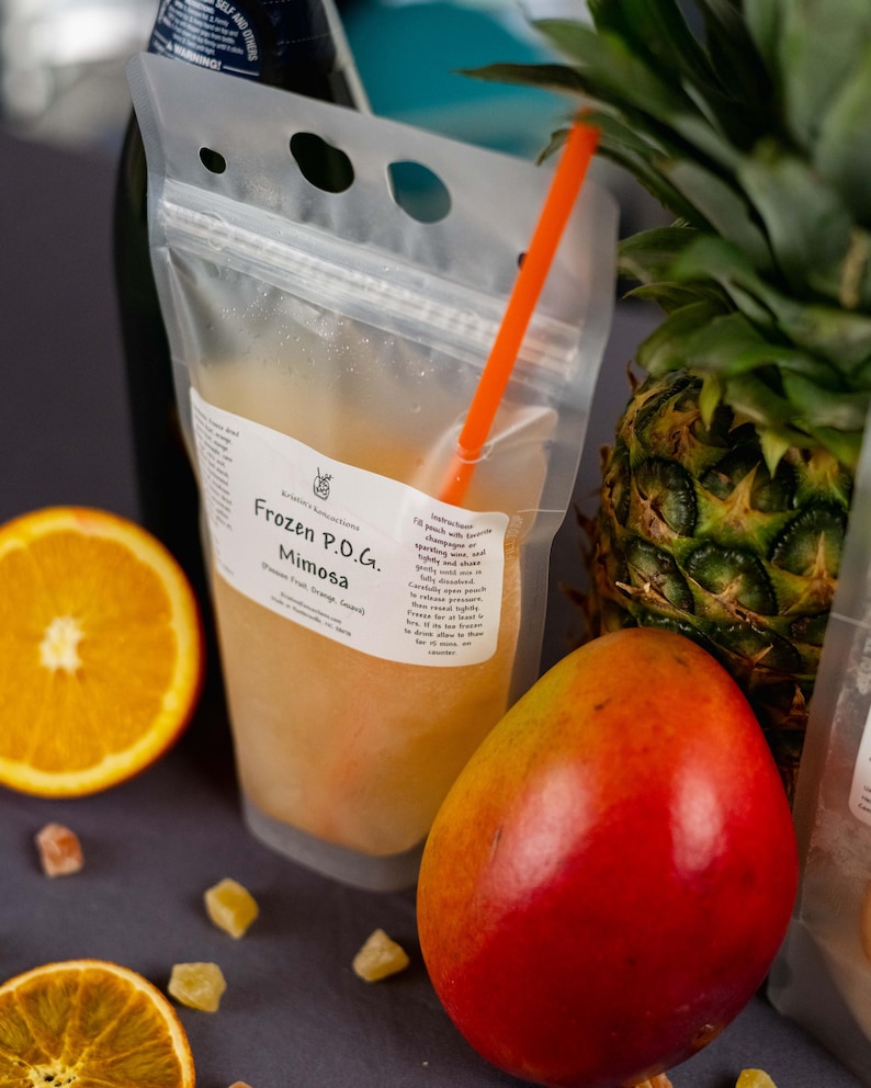 May include: A clear plastic pouch labeled "Frozen P.O.G. Mimosa" with an orange straw, next to a sliced orange, a mango, and a pineapple. The pouch contains a yellow-orange liquid. Small pieces of candied fruit are scattered around the pouch.