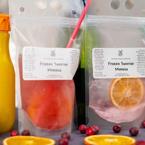 May include: A vibrant image showcasing a mimosa cocktail kit. It includes a bottle of orange juice, a pouch with a red liquid, and a pouch with a dried orange slice. The text "Frozen Sunrise Mimosa" is visible on the pouches. Orange slices and cranberries are scattered around.