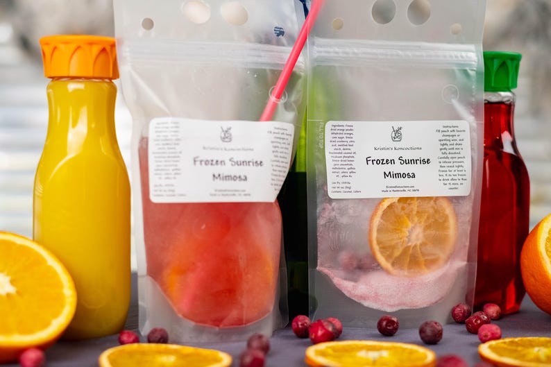 May include: A vibrant image showcasing a mimosa cocktail kit. It includes a bottle of orange juice, a pouch with a red liquid, and a pouch with a dried orange slice. The text "Frozen Sunrise Mimosa" is visible on the pouches. Orange slices and cranberries are scattered around.