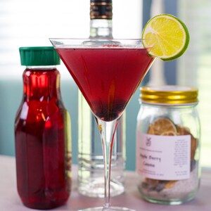 May include: A red cocktail in a martini glass with a lime wedge garnish. The cocktail is garnished with blackberries. There is a bottle of vodka, a bottle of red juice, and a jar of dried fruit in the background.