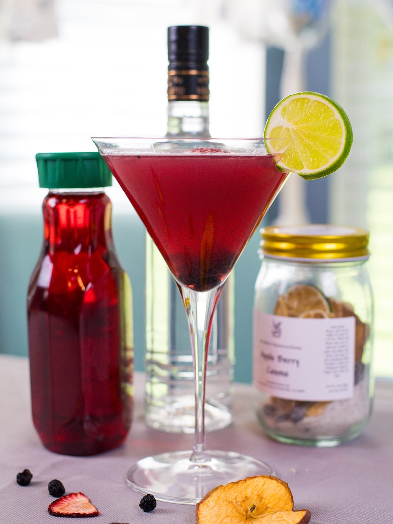 May include: A red cocktail in a martini glass with a lime wedge garnish. The cocktail is garnished with blackberries. There is a bottle of vodka, a bottle of red juice, and a jar of dried fruit in the background.