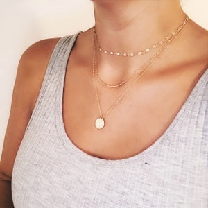 May include: A layered gold necklace set with a delicate chain, a thicker chain with a small round pendant, and a thin bar chain. The necklace is worn by a person with a light grey tank top.