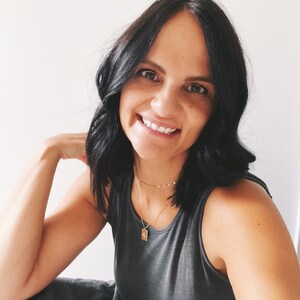 May include: A woman with long black hair is smiling and wearing a grey tank top and a gold necklace with a small pendant.