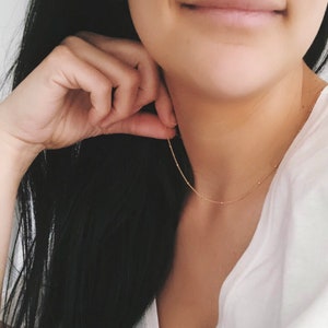Peut inclure: Un collier délicat en chaîne dorée avec de petites perles rondes. Le collier est porté par une personne aux longs cheveux noirs.