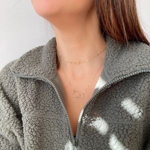 May include: A layered gold necklace set. The top necklace is a delicate chain, and the lower necklace features two interlocking circles. The necklaces are worn against a grey fleece jacket.