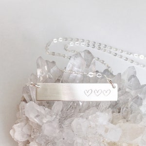 May include: A silver necklace with a bar pendant engraved with three hearts. The necklace is displayed on a bed of white crystals.