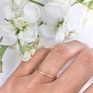May include: A thin gold band ring worn on a finger. The ring is simple and delicate, with a smooth, polished finish.