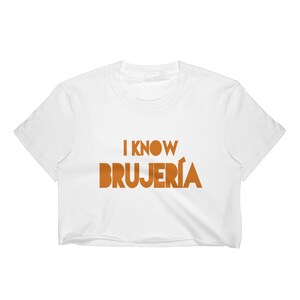 Puede incluir: Top blanco corto de manga corta con el texto "I know BRUJERIA" en naranja.