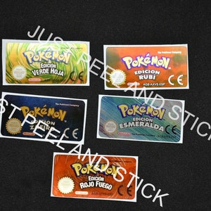 May include: Five different Pokemon game cartridge stickers. The stickers are yellow, orange, blue, green, and red. Each sticker has the Pokemon logo and the text "Edicion" followed by the name of the game in Spanish. The stickers are all CE certified.