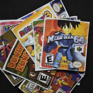 Könnte beinhalten: Ein Stapel von Videospielkassetten für die Nintendo 64 Konsole. Die Kassetten sind für die Spiele Mega Man 64, Donkey Kong 64 und Mario Kart 64. Die Kassetten sind alle von der ESRB mit "E" für Jedermann bewertet.