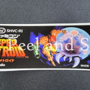 May include: A black and white Super Nintendo game cartridge label for Super Metroid. The label features a red and orange space suit character with a gun, a blue and white planet, and the text "Super Metroid" in red and yellow letters. The text "Nintendo SHVC-RI" and "Super Famicom" is also visible on the label.