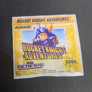 May include: A yellow and blue video game cartridge for the Sega Genesis console. The cartridge features a cartoon character wearing a blue suit of armour and a helmet. The text on the cartridge reads "Rocket Knight Adventures" and "Konami".