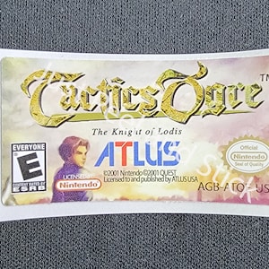 Puede incluir: Una etiqueta de cartucho de juego blanca para el videojuego Game Boy Advance "Tactics Ogre: The Knight of Lodis". La etiqueta presenta un título en dorado, un retrato de personaje y el desarrollador del juego, Atlus. La etiqueta también incluye la clasificación ESRB "Todos" y el sello de calidad de Nintendo.