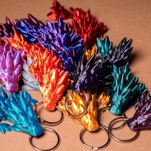Könnte beinhalten: Eine Sammlung von bunten, 3D-gedruckten Drachen-Schlüsselanhängern. Jeder Schlüsselanhänger zeigt einen Drachen mit einem stacheligen, strukturierten Design. Die Drachen sind in verschiedenen Farben, darunter Lila, Blau, Grün, Gold und Rot.