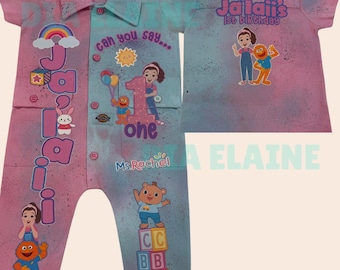 May include: A pink and blue jumpsuit with a collared shirt design. The jumpsuit features the name "Jailaii" and the text "1st Birthday". It has cartoon characters, the number "1", and the text "one".