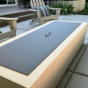 May include: A rectangular outdoor fire pit with a dark gray metal lid and a silver handle. The fire pit is made of light-colored stone and is surrounded by patio furniture, including wooden chairs and green cushions.