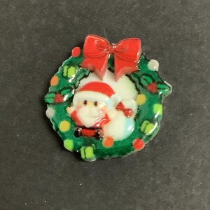 May include: A festive Christmas pin. It features a green wreath with red berries and a red bow. Inside the wreath is a Santa Claus figure. The pin is made of a glossy material.