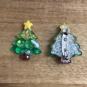 May include: Two Christmas tree pins made of green translucent material. The front view shows a yellow star topper, small colourful dots, and a red base. The back view shows a silver metal pin mechanism attached to a glittery, textured surface.