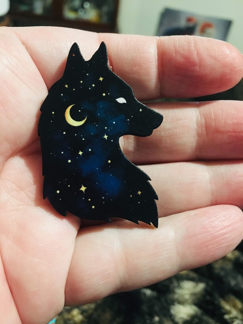 Pu&ograve; includere: Una silhouette nera di una testa di lupo con una luna crescente e stelle al suo interno. La testa di lupo &egrave; fatta di legno e ha uno sfondo blu scuro e nero.