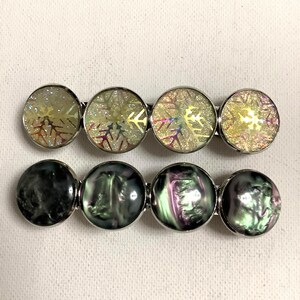 May include: Two hair barrettes with silver-toned metal settings. The top barrette features four circular cabochons with a gold-toned glittery snowflake pattern. The bottom barrette displays four circular cabochons with a dark, iridescent, swirling finish.