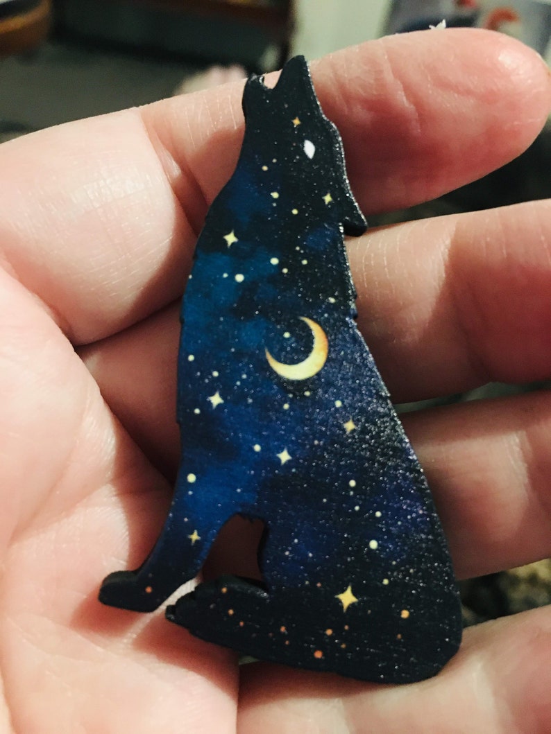 Pu&ograve; includere: Un ritaglio di legno di un lupo che ulula alla luna, con uno sfondo di cielo notturno stellato. Il lupo &egrave; blu scuro, la luna &egrave; gialla e le stelle sono bianche.