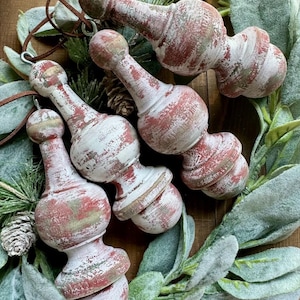 May include: Four distressed, red and white wooden ornaments with a vintage aesthetic. Each ornament has a unique shape and is strung with a brown cord. The ornaments are arranged on a bed of green and gray foliage. The text "Marigold Design Co." is visible.