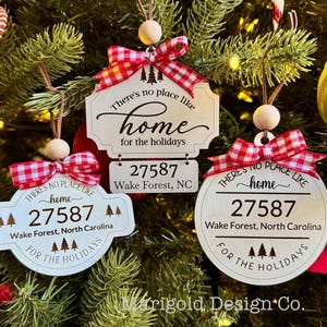 May include: Three white wooden Christmas ornaments with red and white gingham ribbon bows. Each ornament features the text "There's no place like home" with a gold script font. The ornaments also include the text "for the holidays" and the zip code "27587" with the city and state "Wake Forest, North Carolina".
