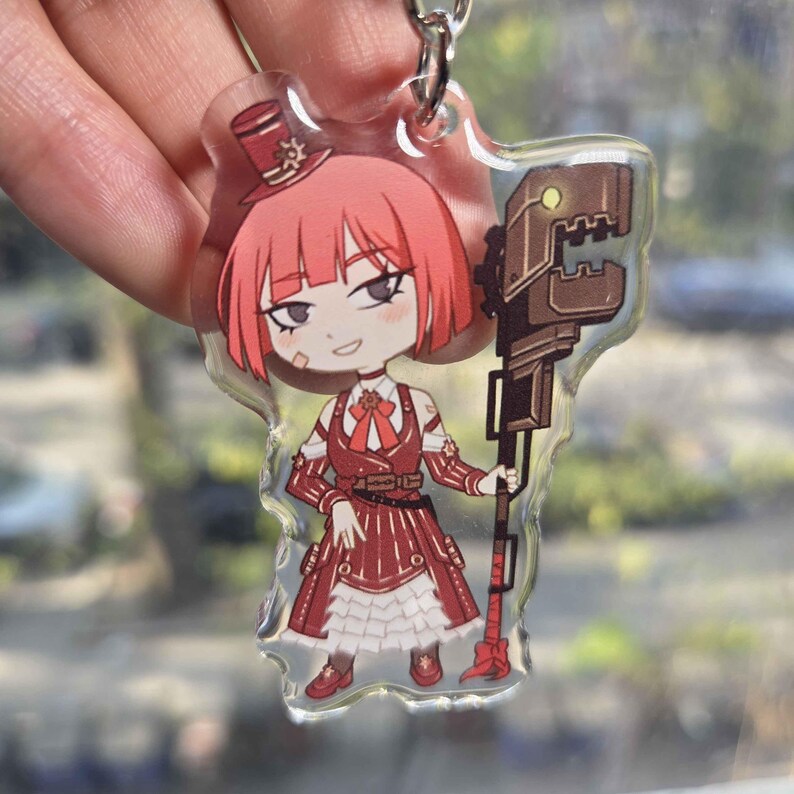 May include: A clear acrylic keychain featuring a cartoon character with red hair and a red outfit, holding a large brown weapon. The character wears a top hat and has a small bandage on their face. The keychain is attached to a silver metal ring.