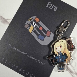 May include: A clear acrylic keychain featuring a cartoon character with blonde hair, red eyes, and a blue jacket. The keychain is attached to a silver clasp. A black card with the name "Ezra" and the text "I'm the talented sidekick, Ezra!" is in the background.