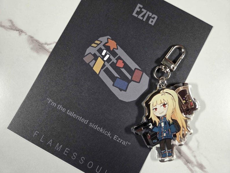 May include: A clear acrylic keychain featuring a cartoon character with blonde hair, red eyes, and a blue jacket. The keychain is attached to a silver clasp. A black card with the name "Ezra" and the text "I'm the talented sidekick, Ezra!" is in the background.