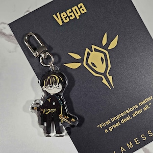 May include: A clear acrylic keychain featuring a cartoon character with glasses and a sword. The keychain is attached to a silver clasp. A black card with gold text that reads "Vespa" and "First impressions matter a great deal, after all." is behind the keychain.