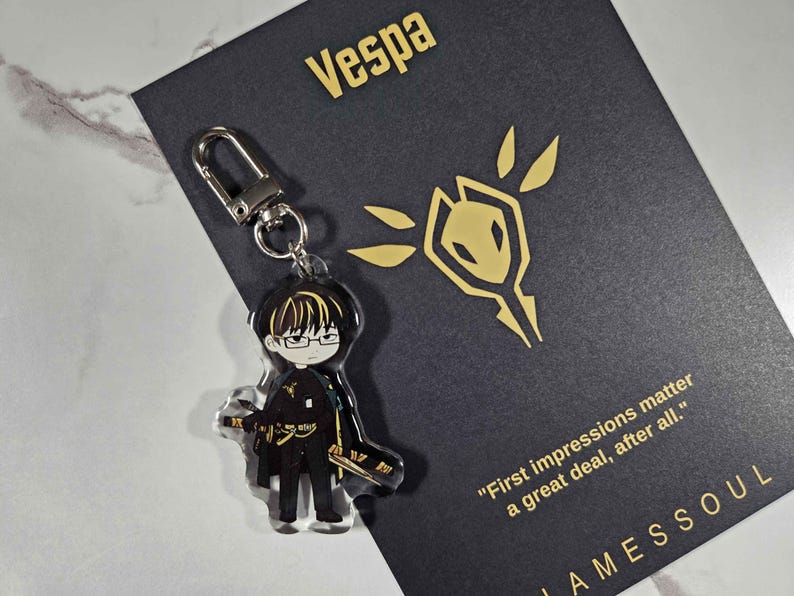 May include: A clear acrylic keychain featuring a cartoon character with glasses and a sword. The keychain is attached to a silver clasp. A black card with gold text that reads "Vespa" and "First impressions matter a great deal, after all." is behind the keychain.