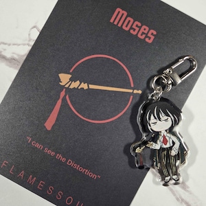 May include: A keychain featuring an anime-style character with black hair, a white shirt, and a red tie. The keychain is attached to a black card with the name "Moses" and the text "I can see the Distortion" in red.