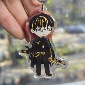 May include: Clear acrylic keychain featuring a cartoon character with black hair, glasses, and a black outfit with gold accents. The character is holding two swords. The keychain is attached to a silver keyring.
