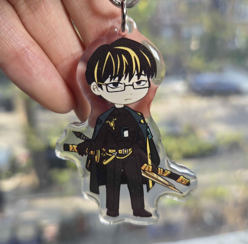 May include: Clear acrylic keychain featuring a cartoon character with black hair, glasses, and a black outfit with gold accents. The character is holding two swords. The keychain is attached to a silver keyring.