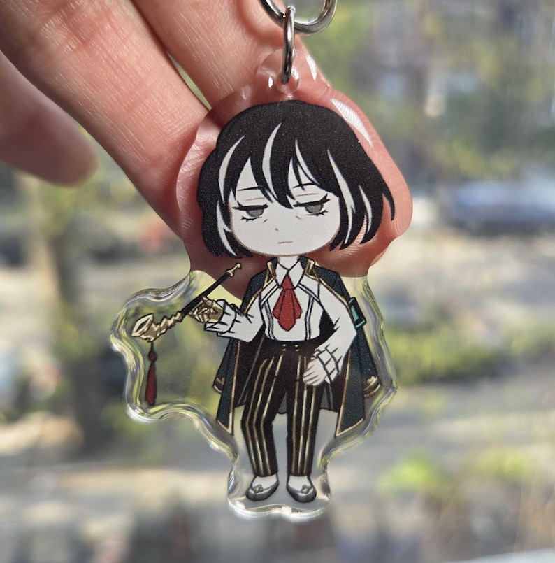 May include: A clear acrylic keychain featuring a cartoon character with black hair, a white shirt, red tie, and black and gold striped pants. The character is holding a decorative wand. The keychain is attached to a silver keyring.