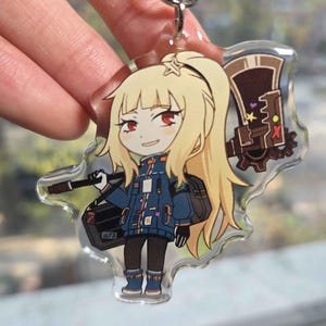 May include: A clear acrylic keychain featuring a cartoon character with blonde hair, red eyes, and a blue jacket. The character is holding a weapon and a black bag. The keychain has a silver metal ring.