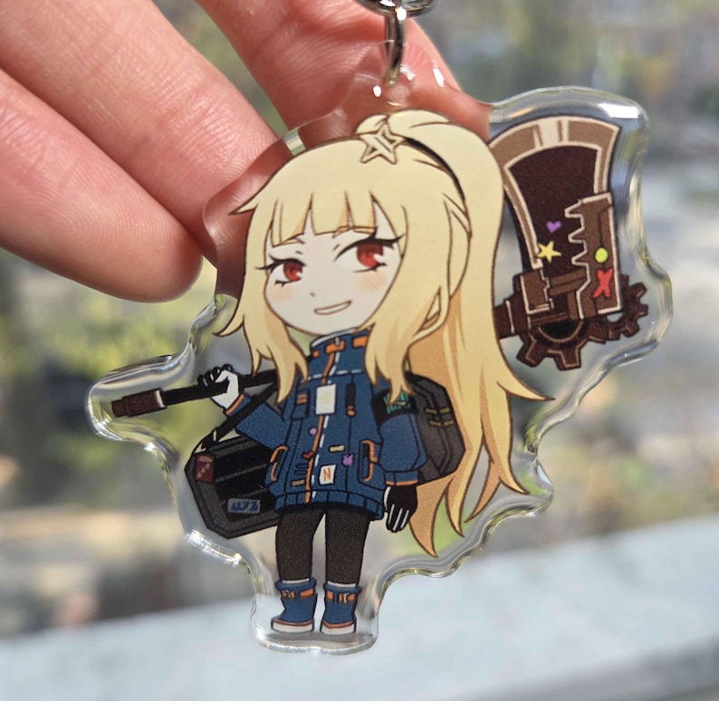 May include: A clear acrylic keychain featuring a cartoon character with blonde hair, red eyes, and a blue jacket. The character is holding a weapon and a black bag. The keychain has a silver metal ring.