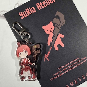 May include: A clear acrylic keychain featuring a red-haired anime-style character in a red and white dress, holding a brown tool. The keychain is attached to a silver clasp. A black card with the text "YuRia Atelier" and a quote is in the background.