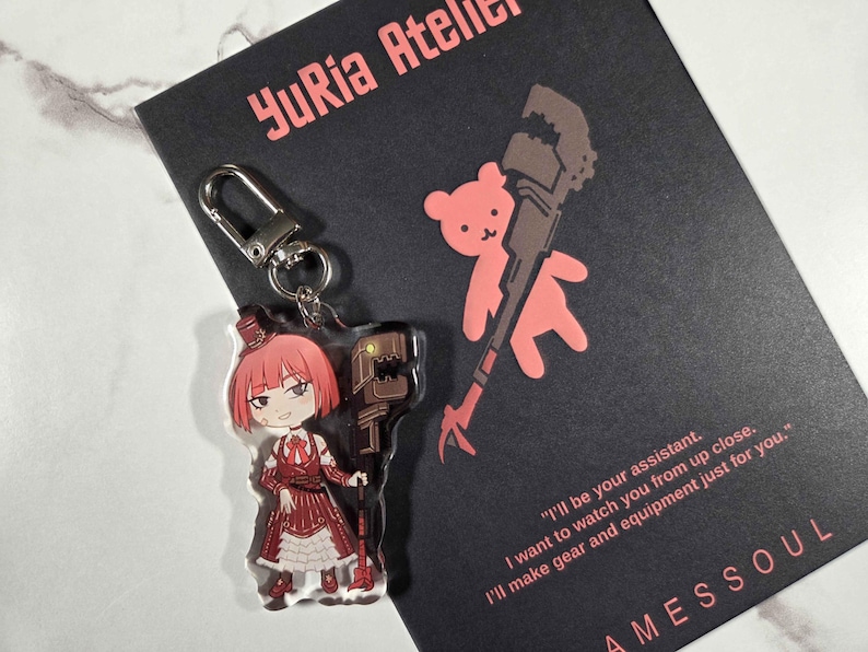 May include: A clear acrylic keychain featuring a red-haired anime-style character in a red and white dress, holding a brown tool. The keychain is attached to a silver clasp. A black card with the text "YuRia Atelier" and a quote is in the background.