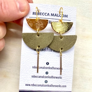 May include: Gold-tone and olive green leather earrings. The earrings feature a half-circle design with a gold-tone top and olive green leather bottom, connected by a gold-tone bar. The earrings are displayed on a white card with the text "Rebecca Malcom Leatherworks".