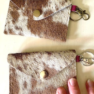 May include: Two rectangular pouches crafted from brown and white cowhide leather, each featuring a triangular flap secured with a brass-toned snap button. A small pink leather tab connects a metal lobster clasp keychain to the side of each pouch.