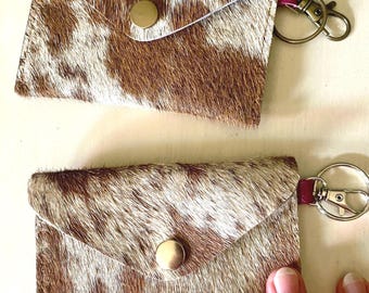 May include: Two rectangular pouches crafted from brown and white cowhide leather, each featuring a triangular flap secured with a brass-toned snap button. A small pink leather tab connects a metal lobster clasp keychain to the side of each pouch.